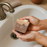 Person holding handcrafted latte soap while washing hands at a bathroom sink