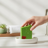 Hand holding a vibrant green Holiday Grump soap bar with a grinch face and red heart on a soap dish