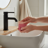 Person holding Sweet Grace inspired silk & goat milk soap bar under running water in a stylish bathroom